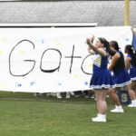 Cheerleaders in blue uniforms stand in front of a large banner that reads "Go t," waving their arms during a sports event on a grassy field.