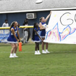 Three cheerleaders in blue uniforms hold a colorful banner on a grassy field, preparing for a sports event. People stand in the background near a blue building.