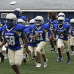 A youth football team in blue and white uniforms runs onto the field, wearing helmets and pads.