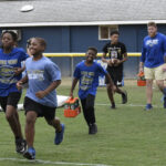 Children and teenagers run and smile on a grassy field, carrying orange cones, with a building and fence in the background.