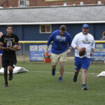 Four people run on a grassy field during a football practice or camp, each holding a football, with banners and a building in the background.