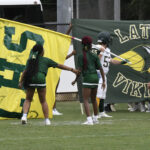 Two cheerleaders hold a large yellow banner as football players in uniform prepare to run onto the field before a game.