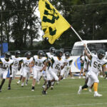 A football team in white uniforms runs onto the field, with one player leading and holding a large yellow flag. Trees and a bus are in the background.