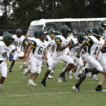 A group of high school football players in white and green uniforms run across a grassy field, with a school bus visible in the background.