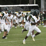 High school football players in white uniforms run onto the field during a game, with cheerleaders and a bus visible in the background.