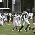 Football players in white and green uniforms run on a grassy field during a game, with cheerleaders and a bus visible in the background.