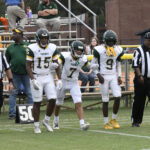 Three high school football players in white uniforms stand on the field near a referee and coaches, with bleachers and spectators visible in the background.