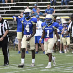 Two football players in blue and gold uniforms walk on the field with a referee, while teammates and spectators watch from the bleachers in the background.
