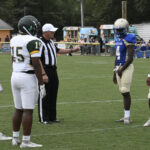 Football players and a referee gather on the field for a coin toss before a game, with food trucks and trees visible in the background.
