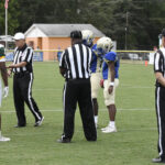 Football players and referees gather on a grassy field, preparing for the start of a game. A scoreboard and small building are visible in the background.