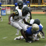High school football players in green and white uniforms tackle a player in blue and gold during a game on a grassy field.