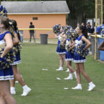 A group of cheerleaders in blue and white uniforms stands on a grassy field, holding pom-poms, performing during an outdoor event.