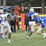 High school football players in blue and white uniforms play on a grassy field; referee, scoreboard marker, and school buses visible in the background.