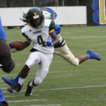 A football player in a white uniform runs with the ball as a defender in blue attempts to tackle him during a game.