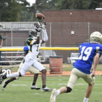 A football player in a white uniform jumps to catch a pass while a player in a blue uniform runs towards him on a field.