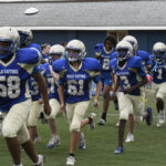 A youth football team in blue and white uniforms runs onto a field, wearing helmets and pads, appearing energetic and focused.