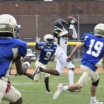 Football player in white catches a pass while surrounded by opponents in blue jerseys on a grassy field during a daytime game.