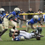 High school football players in blue and white uniforms tackle a player in green and white on a grassy field during a game.