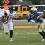 Two high school football players in action on the field, one running with the ball as another prepares to tackle. School bus in background.