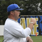 A coach wearing a blue cap and white shirt stands on a sports field, holding a whistle, with a large sign and trees in the background.