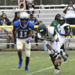 Two high school football players chase a loose football on the field as spectators watch from the bleachers in the background.