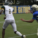 A football player in white runs with the ball while a player in blue tries to catch up on a grassy field.