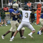 Two high school football players in action on the field, with one in blue and the other in white, as spectators watch from the bleachers.