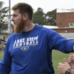 A man wearing a blue "Lake View Football" shirt stands outdoors, looking to the side, with trees and buildings visible in the background.