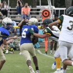 Youth football players in blue and white uniforms run and block during a game, with spectators watching from bleachers in the background.