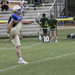 Football player in blue and gold uniform executing a high kick on the field, with cheerleaders and a scoreboard in the background.