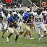 A high school football game with players in blue and white uniforms running on the field, while fans watch from the bleachers in the background.