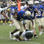 A youth football player in blue runs with the ball while another player in white and green attempts to tackle him during a game.