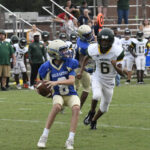 A high school football quarterback in blue prepares to throw as a player in white jersey number 6 chases him on the field.