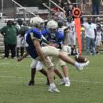 A football player in blue and tan uniform punts the ball during a game, with teammates and opponents nearby and spectators in the background.