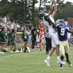 Two high school football players jump to catch the ball during a game, with cheerleaders and spectators watching from the sideline.