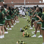 A group of cheerleaders in green uniforms perform routines on a grassy field, with pom-poms on the ground and a sports team visible in the background.