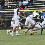 High school football players in white uniforms tackle a player in blue on a grassy field, with spectators watching from bleachers in the background.