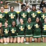A group of cheerleaders in green and yellow uniforms pose for a team photo on a sports field with bleachers and people in the background.