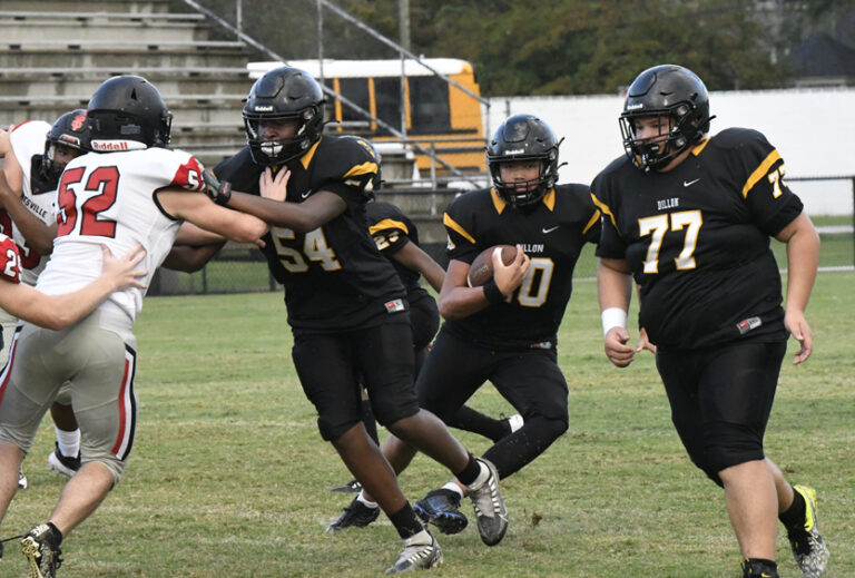 High school football players in black uniforms run a play, with one carrying the ball and teammates blocking defenders on a grassy field.