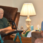 Two elderly men sit in recliners in a cozy living room with a lit table lamp and a walker between them.