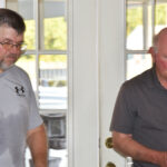 Two men standing indoors near glass doors, both wearing casual shirts, one with glasses.