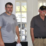 Two men standing indoors; one wears a sweat-stained gray t-shirt, the other a dark polo and cap. Both look towards the camera.