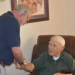 elderly man sitting in a chair shakes hands with another man standing beside him in a warmly lit room.