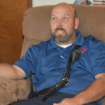 A man with a beard sits on a recliner chair, wearing a blue shirt and a black shoulder strap, gesturing with his hand.