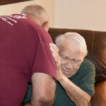 older man sitting on a couch is being comforted by another person who is giving him a hug.