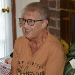 older woman with short hair and glasses, wearing a tan shirt, sits indoors near a brick wall and smiles.