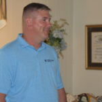 Man in a light blue polo shirt standing indoors, with framed picture and flowers in the background.