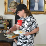 A young person holding a red cup and plate of food stands next to an adult serving themselves at a food table in a home.