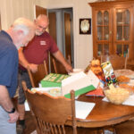Two men look at food on a dining table with pizza boxes, chips, and snacks in a cozy dining room with wooden furniture.