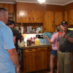 Several people gather in a wooden kitchen, chatting and preparing drinks. Bottled drinks and snacks are on the counter, creating a casual, social atmosphere.